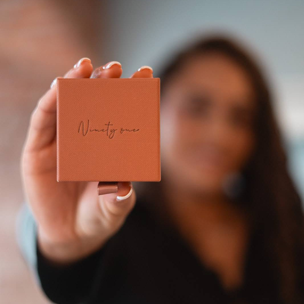 Person holding a small orange box with 'Ninetyone' branding, blurred background