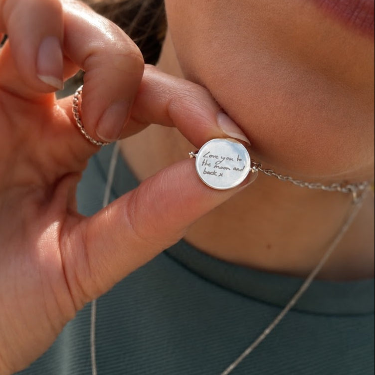 A close-up lifestyle shot of a person wearing a delicate silver chain necklace with a small oval silver disc pendant engraved with the handwritten phrase 'Love you in the storm and rain.' The person is wearing a sage green ribbed top, and the warm sunlight casts a soft shadow on their skin.