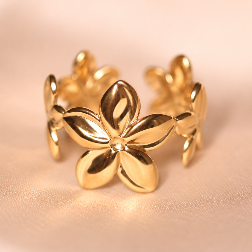 A polished gold-tone ring featuring a central five-petal flower design with smaller matching flower accents along the band, displayed on a soft beige fabric background.