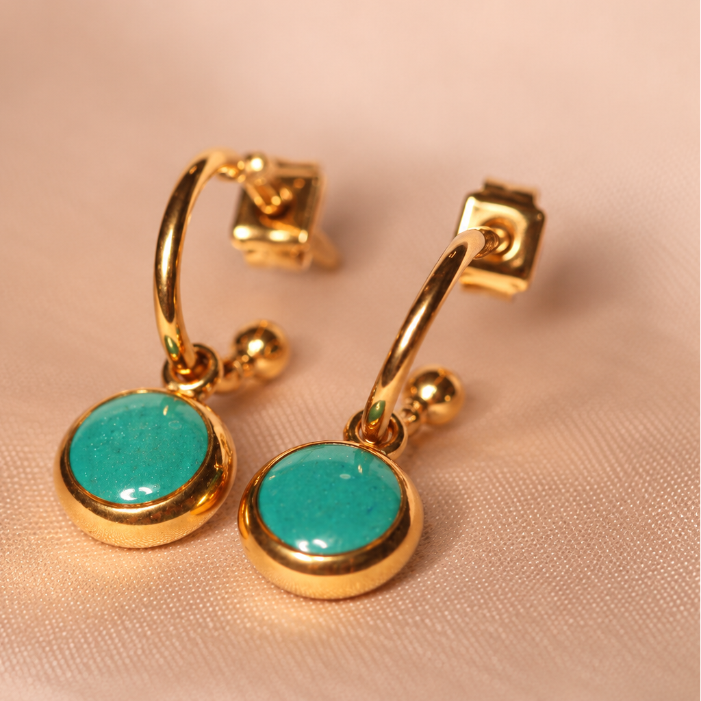 A pair of gold huggie hoop earrings with round turquoise cabochon stones, displayed on a soft beige fabric background.