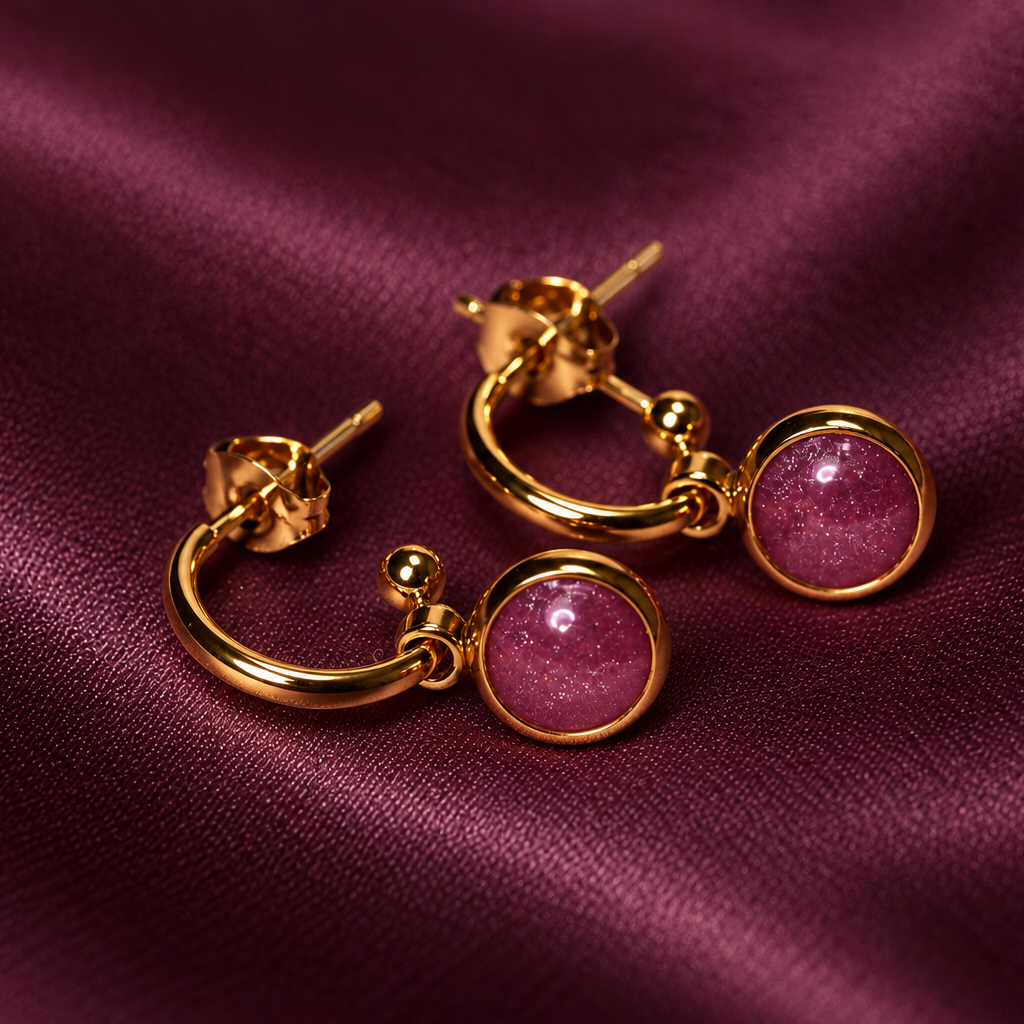 A pair of gold hoop earrings with round pink gemstone drops, displayed on a draped dark purple satin fabric background.