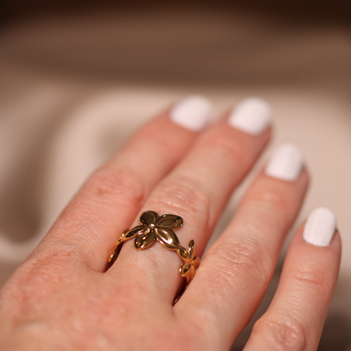 A close-up of a hand with white manicured nails wearing a gold ring shaped like a stylized clover or leaf cluster, set against a soft beige fabric background.