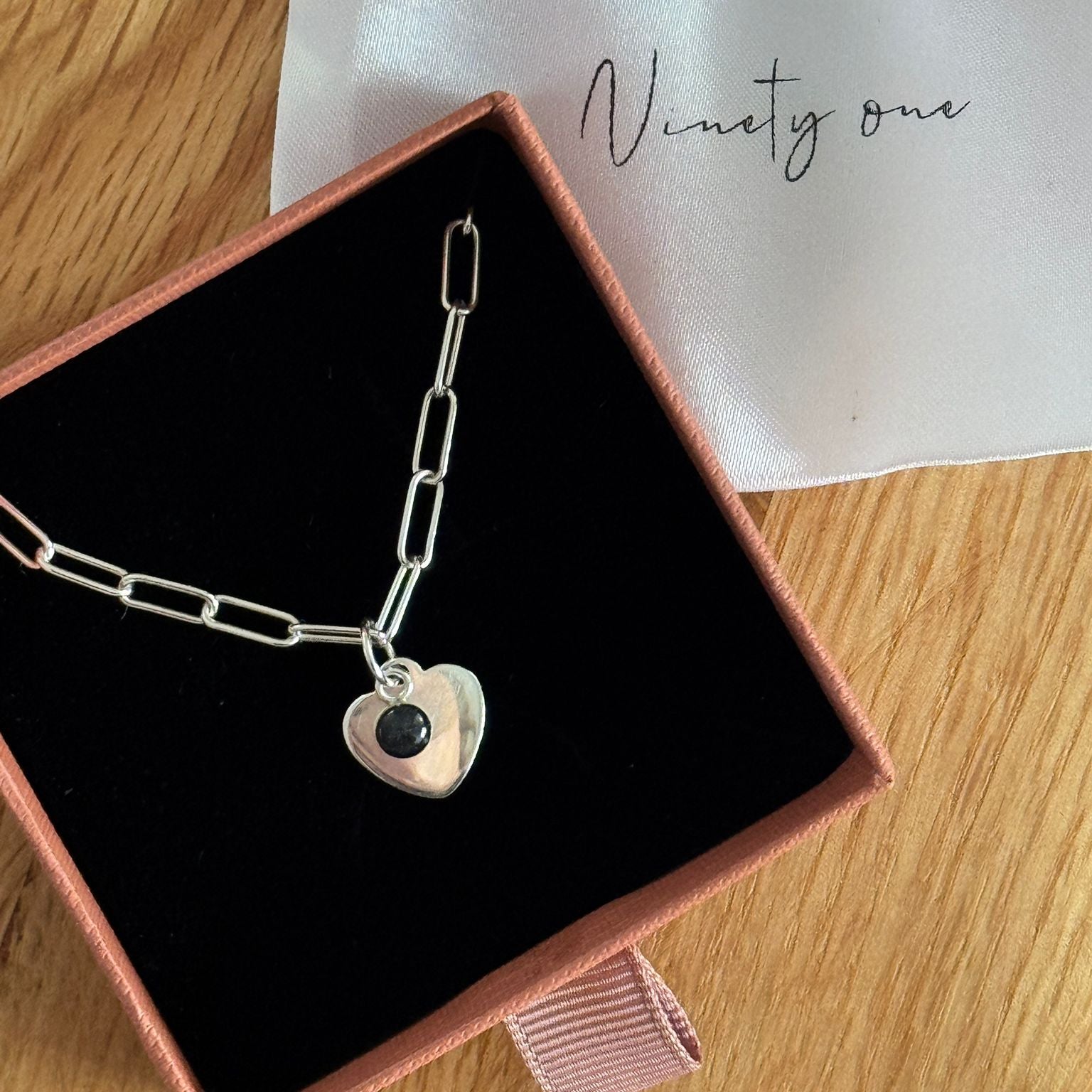 Silver bracelet with a heart-shaped charm in a black box on a wooden surface.