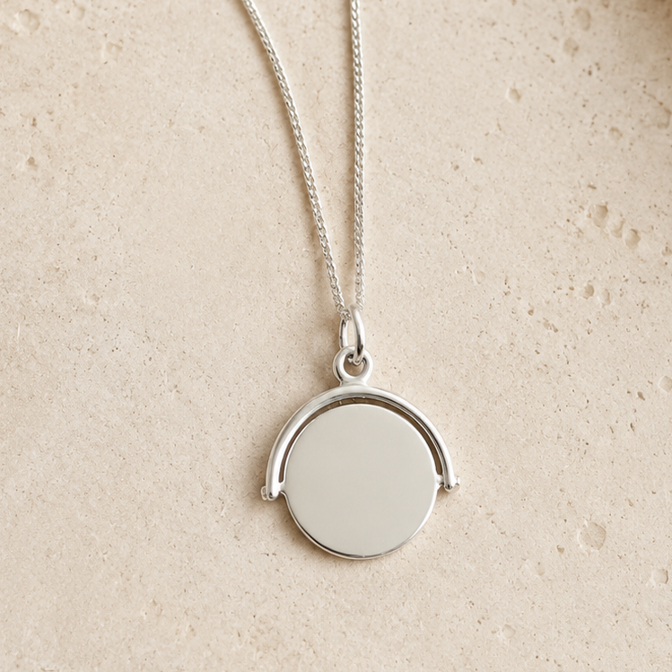 A silver necklace with a round disc pendant and an open circular arch detail, displayed on a textured beige stone surface.