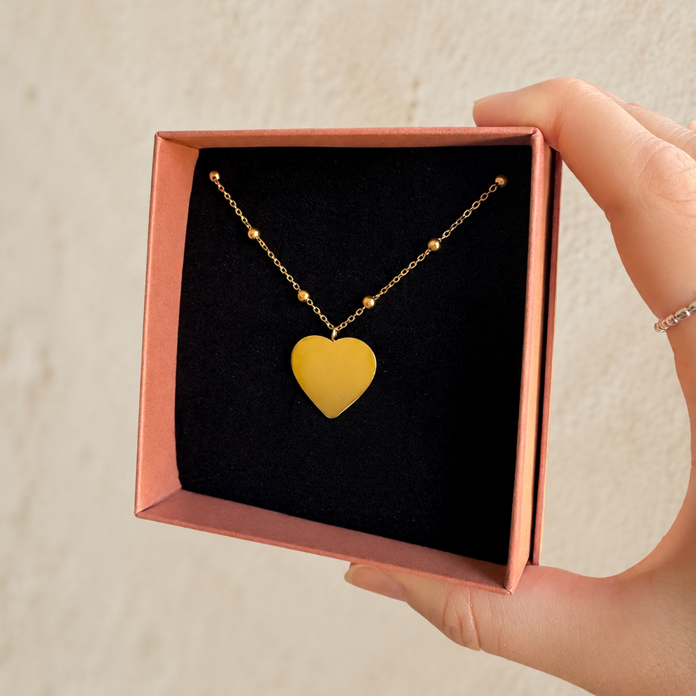 A gold chain necklace with a gold heart pendant displayed inside an open pink gift box held by a hand against a light textured wall.