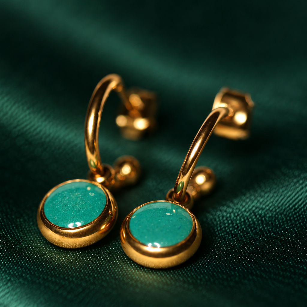 A pair of gold huggie hoop earrings with round turquoise-blue cabochon stones, displayed on a dark green satin fabric background.