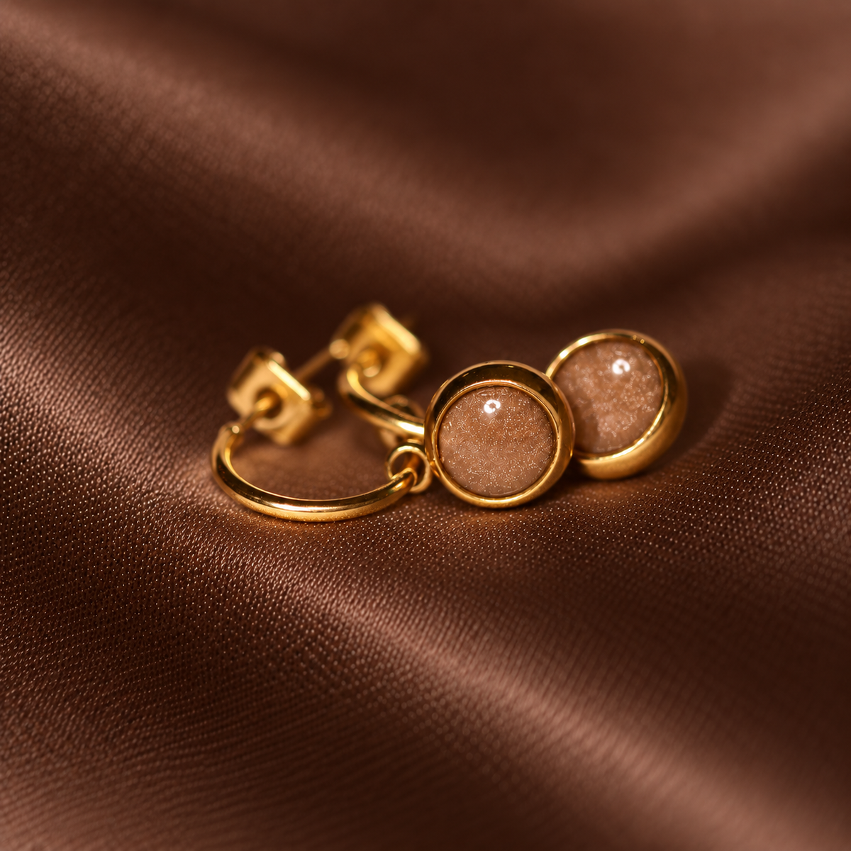 A pair of gold hoop earrings with round, textured brown stone inlays, displayed on a draped brown satin fabric.