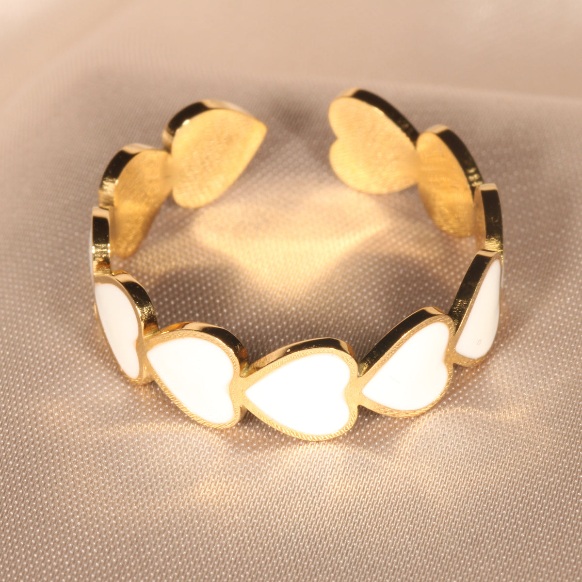 A gold-plated cuff bracelet featuring a series of white enamel heart-shaped links, displayed on a soft beige fabric background.