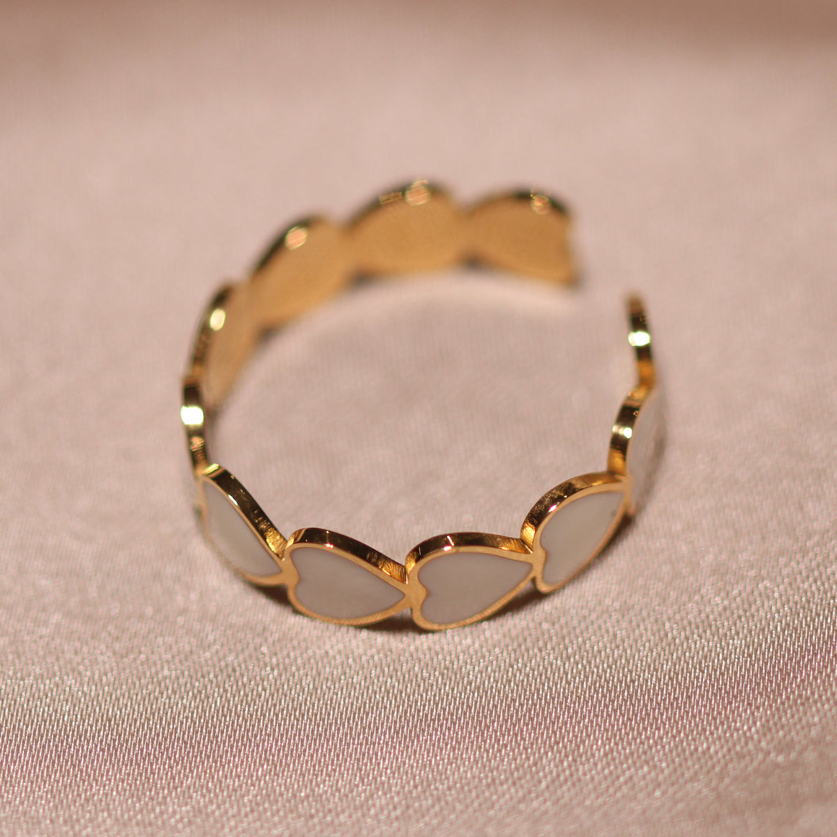 A gold-plated open ring band featuring a series of small, scalloped oval inlays with a pale, iridescent finish, resting on a soft, draped beige fabric.