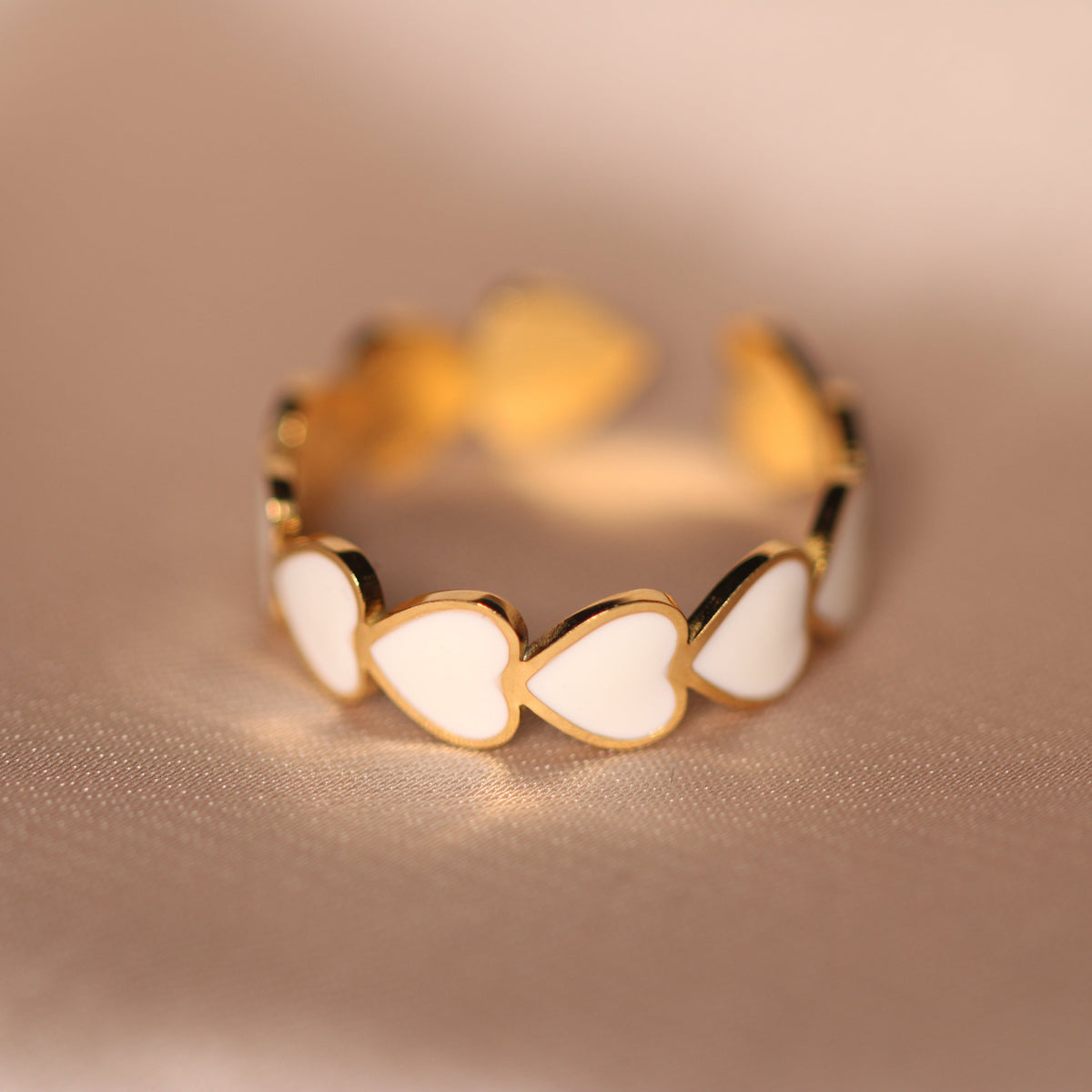 A gold adjustable ring featuring a series of white heart-shaped enamel inlays, photographed on a soft beige satin background.