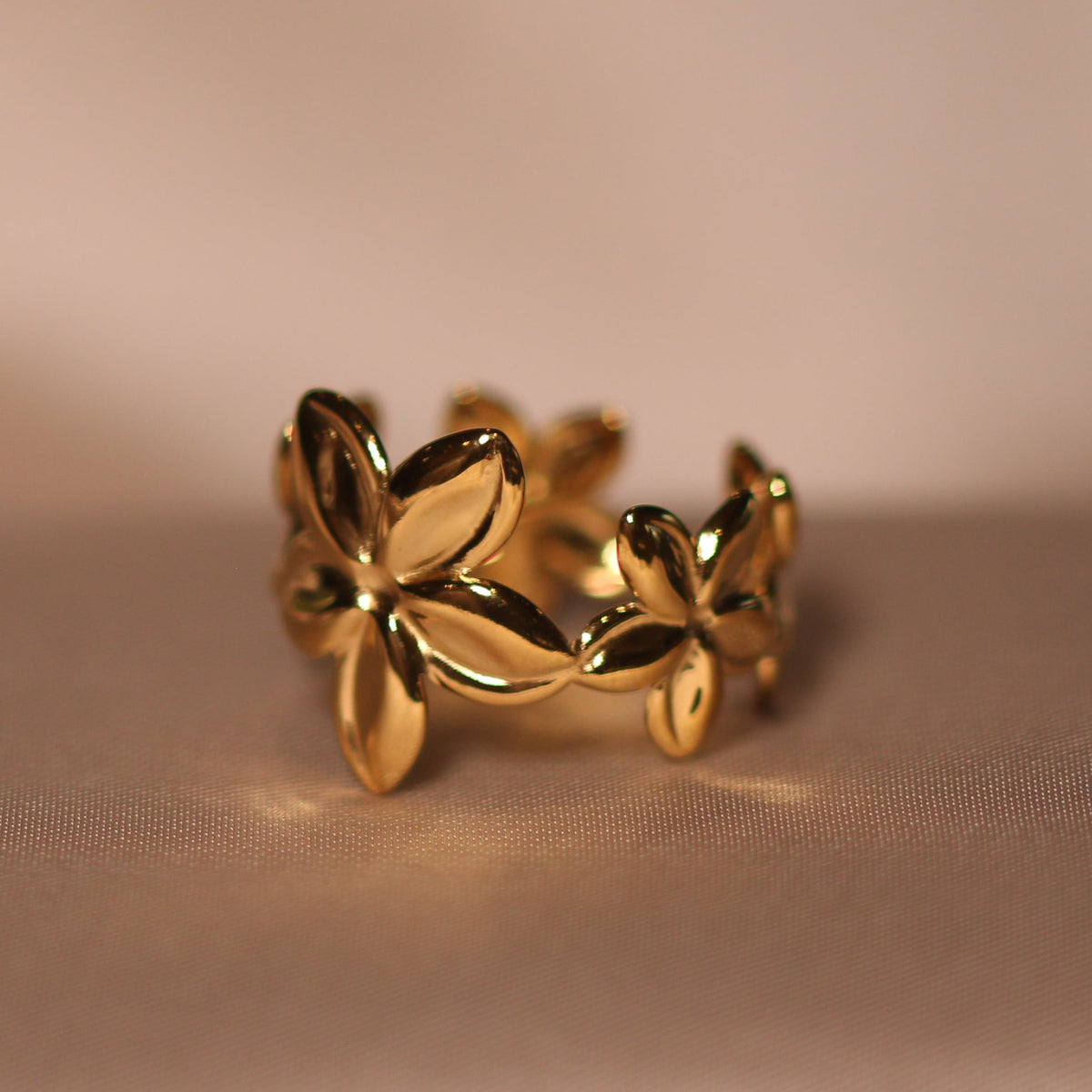 A gold ring with a stylized floral or leaf-like openwork design, resting on draped beige satin fabric.