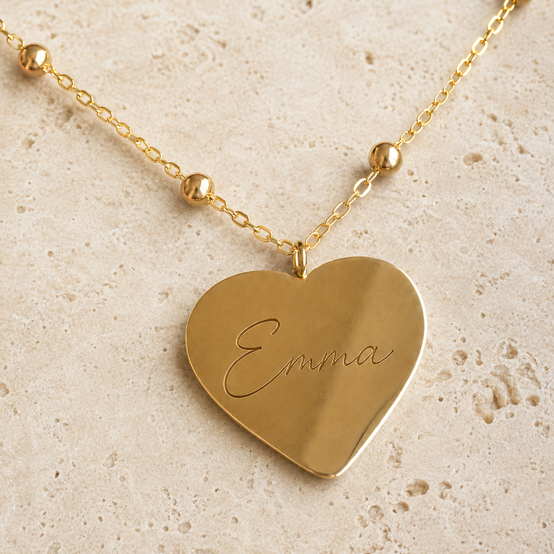 A gold heart-shaped pendant necklace engraved with the name 'Emma', displayed on a textured beige stone surface.