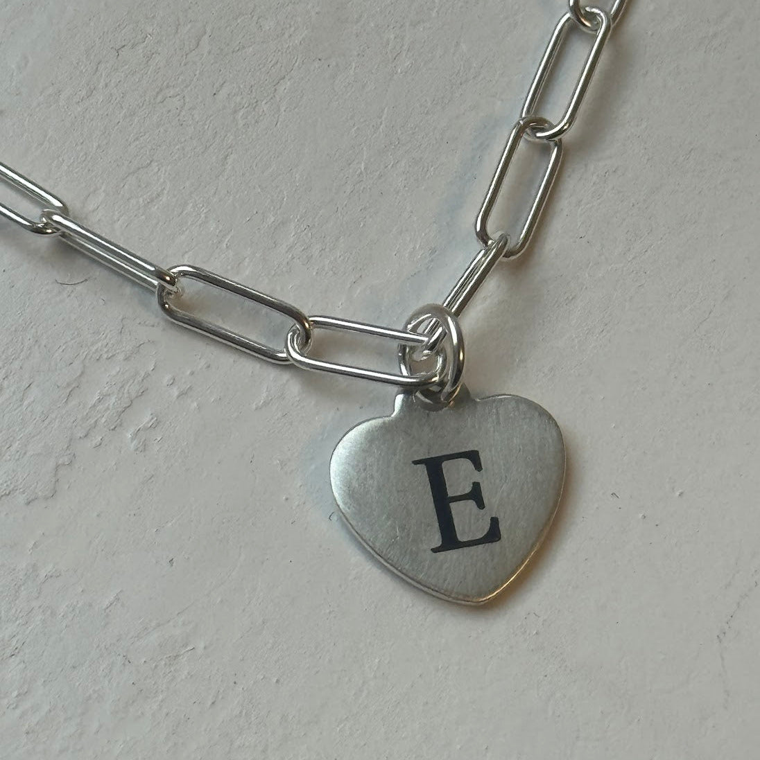 Silver chain bracelet with a heart-shaped charm featuring the letter 'E' on a light gray background.