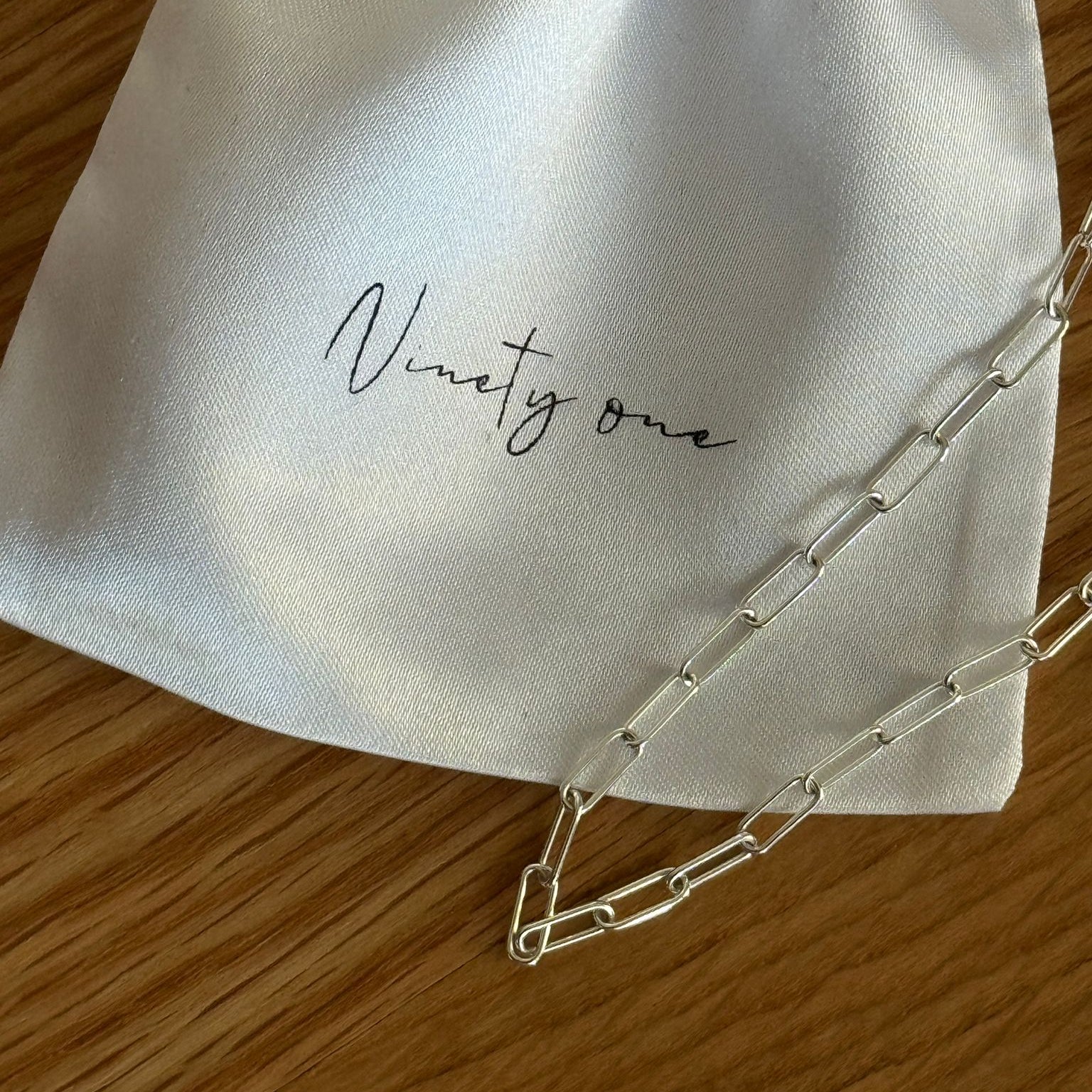 Silver chain on a white drawstring bag with 'Ninety one' embroidery on a wooden surface